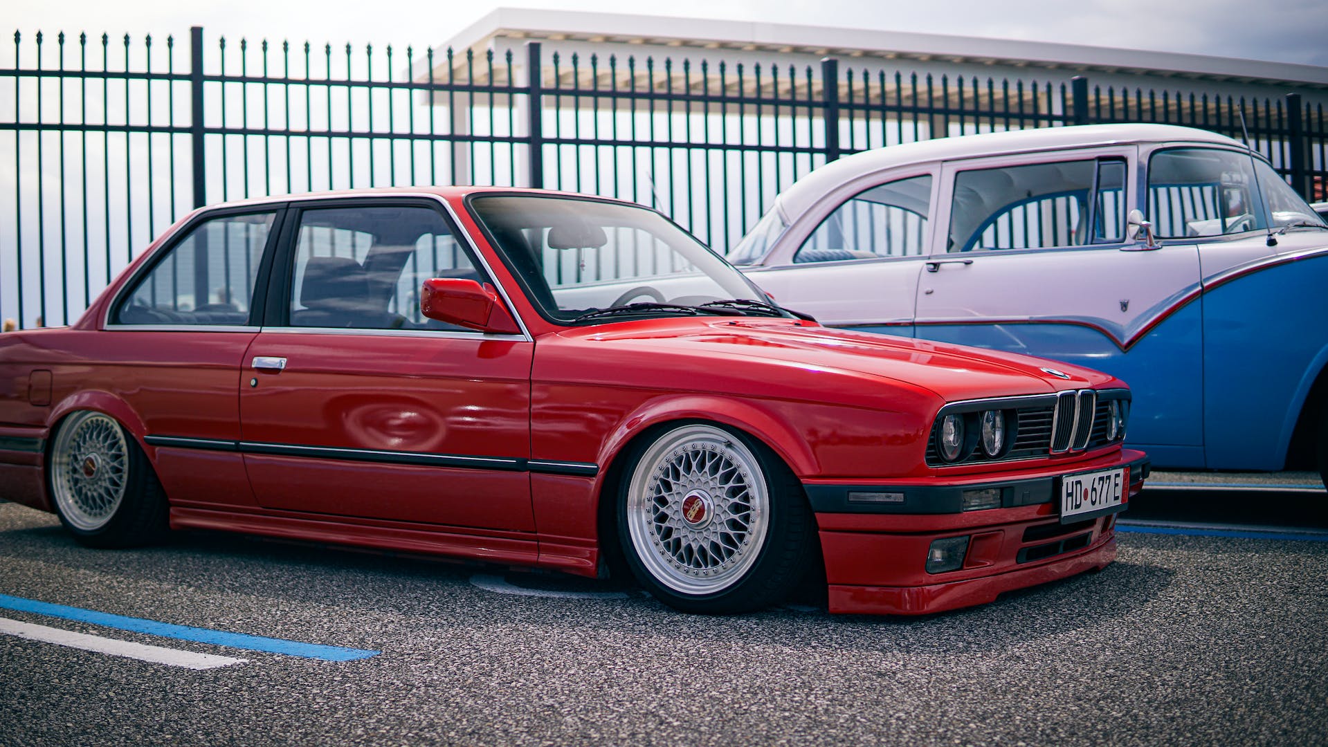 A classic red BMW E30 model with distinctive BBS rims is parked in front of a blue and white vintage car, both vehicles set against a backdrop of a metal fence and overcast sky.