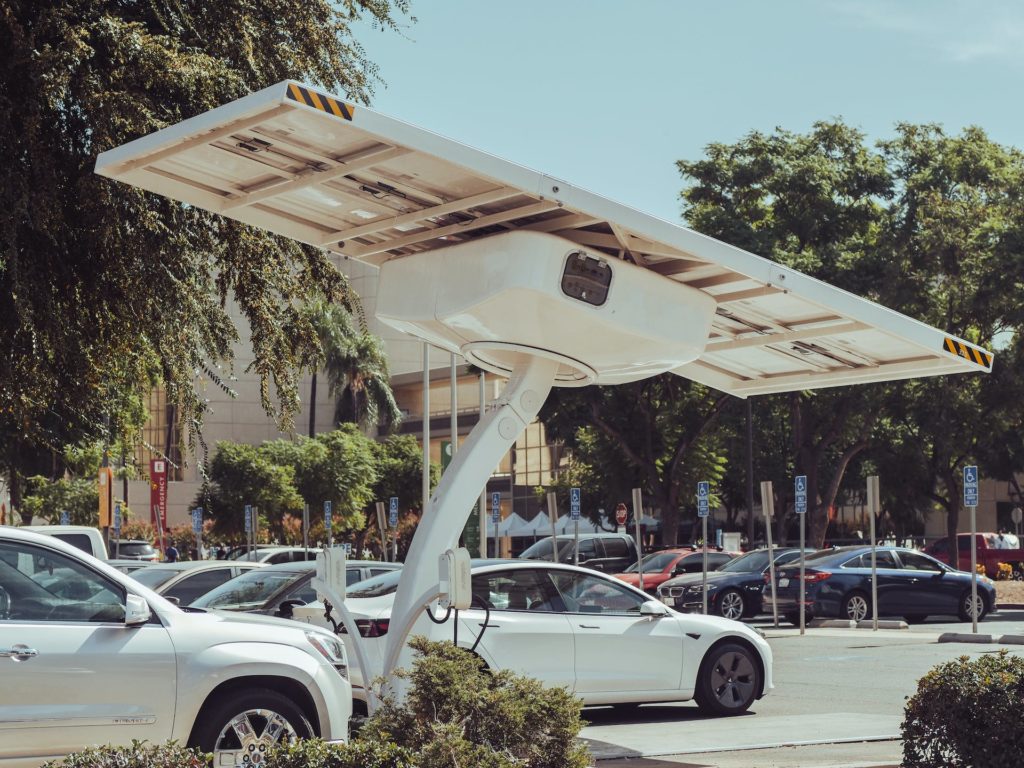 A white electric vehicle is parked under a futuristic solar panel canopy in an urban parking lot, highlighting a blend of modern clean energy solutions and urban transportation.