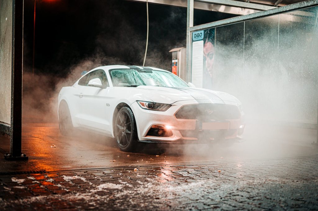 A white Ford Mustang GT with its headlights on is partially shrouded in steam or mist, giving the scene a dramatic look. The car is in a car wash bay, with water droplets visible on the ground and car wash equipment in the background.